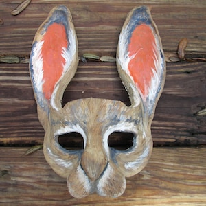 May include: A hand-carved wooden rabbit mask with large ears. The inside of the ears are painted orange, with black and white accents. The mask has eye and nose openings, and is set against a rustic wooden background.