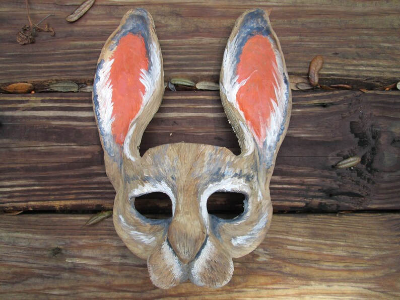 May include: A hand-carved wooden rabbit mask with large ears. The inside of the ears are painted orange, with black and white accents. The mask has eye and nose openings, and is set against a rustic wooden background.