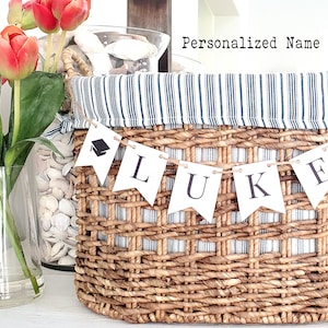 May include: A woven wicker basket with a blue and white striped fabric lining. A banner spelling "LUKE" with graduation cap icons hangs across the front. The text "Personalized Name Banner" is above the basket.