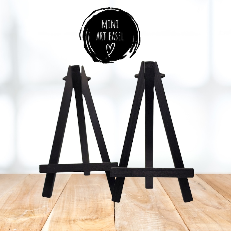Op de afbeelding: Twee kleine zwarte houten mini-schildersezels op een licht houten oppervlak. De ezels zijn klein en eenvoudig, perfect voor het weergeven van kleine kunstwerken of foto's.