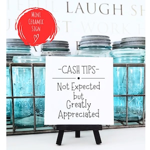 May include: White ceramic sign with black text that reads "CASH TIPS - Not Expected but Greatly Appreciated". The sign is on a small black easel and is sitting on a table with blue glass jars.
