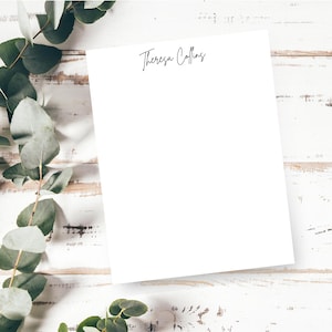 May include: A blank white sheet of stationery with the name "Theresa Collins" in elegant black script at the top. The paper is positioned on a distressed white wooden surface, with green eucalyptus leaves in the background.