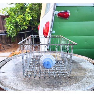 May include: A vintage-style wire basket with a blue and white object attached. The basket is made of metal and has a rectangular shape. The background includes a green and white camper and a wooden barrel.