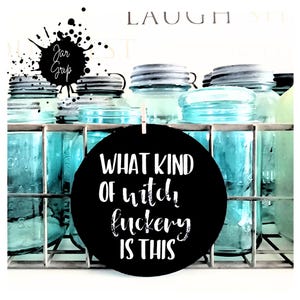 May include: A collection of vintage blue mason jars in a metal wire rack. A black circular sign with the text "WHAT KIND OF witch fuckery IS THIS" is clipped to the jars. The words "Jar Grip" are written in a black splatter design.