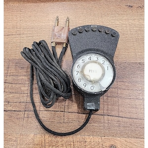 May include: A vintage black telephone with a rotary dial and a cord. The dial features numbers 0-9 and the word "PIXIE". The phone has a series of buttons labeled "RI" and "SEIZE".