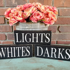 May include: Three black wooden signs with white lettering. The top sign reads "LIGHTS", the middle sign reads "WHITES", and the bottom sign reads "DARKS".
