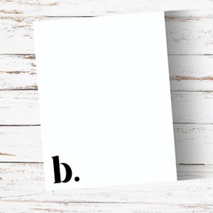 May include: A white rectangular card with the lowercase letter "b" and a period in black at the bottom left corner. The card is positioned at an angle against a white wooden background.