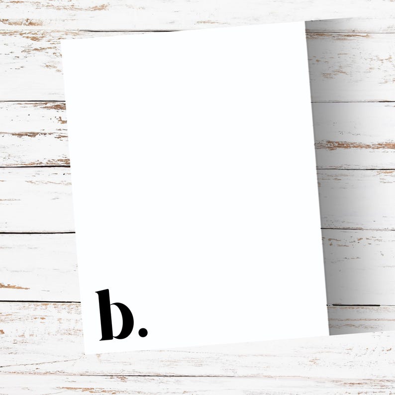 May include: A white rectangular card with the lowercase letter "b" and a period in black at the bottom left corner. The card is positioned at an angle against a white wooden background.
