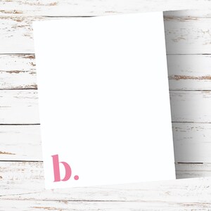 May include: A white sheet of paper with the pink letter "b" and a period in the lower left corner. The paper is set against a white wooden background.