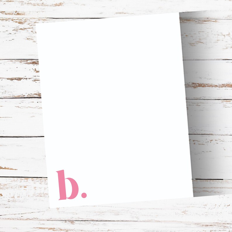 May include: A white sheet of paper with the pink letter "b" and a period in the lower left corner. The paper is set against a white wooden background.