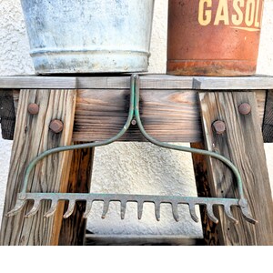 May include: A weathered green metal garden rake head featuring a wide, arched frame and a series of downward-curving tines. The metal shows signs of oxidation and wear, with a distinct patina across the surface of the tool's frame and teeth.