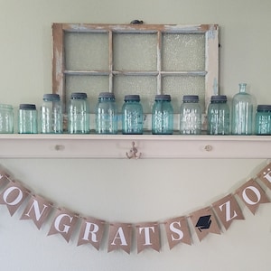May include: A burlap banner with the text "CONGRATS ZOE" in white letters. A graduation cap icon is included in the banner. The banner is hanging on a white shelf with a row of blue glass jars on top.