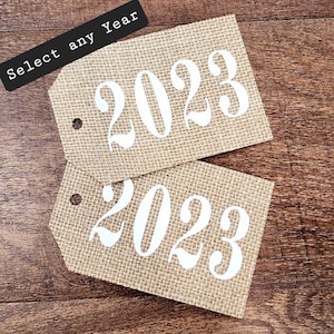 May include: Two burlap tags with the year 2023 printed in white. The tags are on a wooden surface. Select any year.