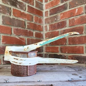 May include: An antique potato ricer with a white and teal handle and a rusty, perforated basket. The kitchen tool is made of metal and sits on a wooden surface against a brick wall.