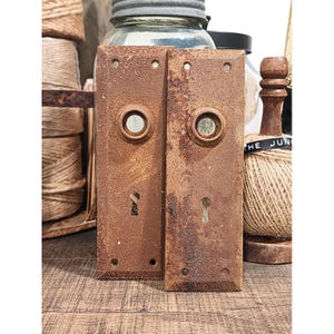 May include: Two antique, rectangular, rusty brown door plates with keyholes and circular knobs. The plates have a weathered appearance, suggesting age and use. The background includes twine, a glass jar, and a wooden spool.