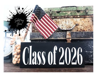May include: A black wooden sign with the text "Class of 2026" in white lettering. An American flag is placed on top of a vintage metal trunk. A jar of round objects and a scale are visible in the background.