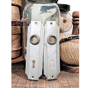 May include: Two vintage, light gray door handle backplates with keyholes and circular accents. The backplates are rectangular with a decorative, stepped design. The background includes jars, twine, and wooden elements, suggesting a rustic or antique theme.