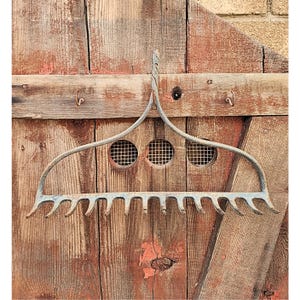 May include: An antique metal garden rake with a curved handle and multiple tines. The rake is painted a faded blue and is hanging on a weathered wooden door with three circular vents.