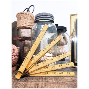 May include: A vintage wooden folding ruler with inch measurements, fanned out on a wooden surface. The ruler is next to glass jars, spools of twine, and a decorative frame, creating a rustic, antique aesthetic.