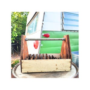 May include: A rustic wooden tool caddy with a handle, featuring a light-colored base and a dark-stained frame. The caddy is filled with small wooden dowels and sits on a wooden barrel. A vintage green and white camper is in the background.