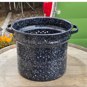 May include: A speckled black and white enamelware colander with two handles. The colander has a cylindrical shape with numerous small holes for draining. The rim and handles are solid black. This kitchenware item is designed for straining pasta, vegetables, or other foods.