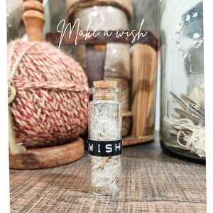 May include: A small glass bottle with a cork stopper, filled with fluffy white and brown seeds. The bottle has a black band that reads "WISH" in white letters. The background includes spools of twine and the text "Make a wish".