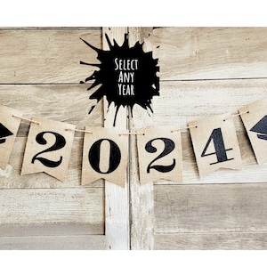 May include: A burlap banner with black graduation caps and the year 2024 printed on it. The banner is strung on a white string and is hanging on a wooden background. The text "Select Any Year" is written in black on a white splatter background.