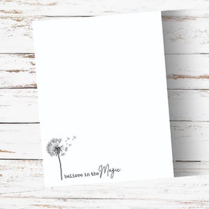 May include: A white card with a black dandelion illustration and the words "believe in the Magic". The dandelion is on the lower left, with the text on the lower right. The card is on a white wooden surface.