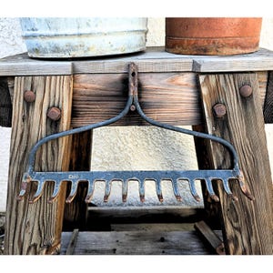 May include: A vintage metal garden rake head featuring a curved support frame and a horizontal bar with eleven downward-pointing tines. The dark grey metal shows signs of oxidation and wear, with the word CHINA stamped clearly on the front bar.