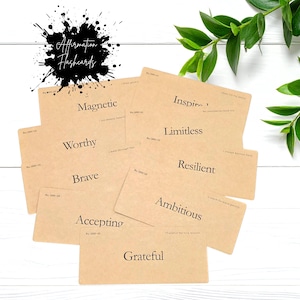 May include: A set of rectangular kraft paper affirmation flashcards featuring black printed text. The cards display individual words including Magnetic, Inspire, Limitless, Worthy, Resilient, Brave, Ambitious, Accepting, and Grateful, with small reference numbers and additional phrases printed on each card.