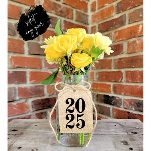 May include: A burlap tag with the year 2025 printed in black, tied with twine around a glass jar filled with yellow flowers. The jar is sitting on a wooden surface.