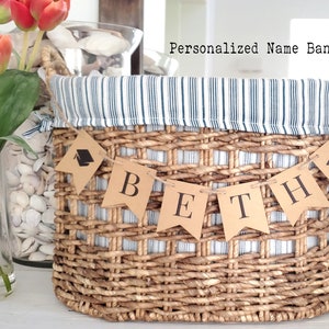 May include: A wicker basket with a blue and white striped liner and a personalized name banner that reads "BETH". The banner is made of brown paper with black letters and graduation caps.