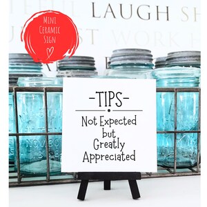 May include: White ceramic sign with black text that reads "TIPS - Not Expected but Greatly Appreciated". The sign is on a small black easel and is in front of a row of blue glass jars.