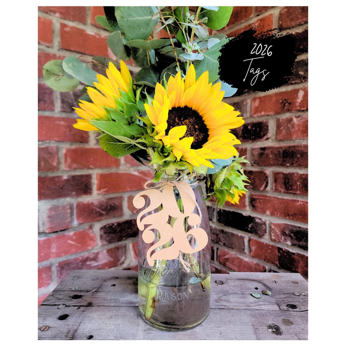 May include: A mason jar vase filled with sunflowers and eucalyptus greenery. A wooden tag with the number "2026" is tied to the vase. The text "2026 Tags" is written in black on a white background.