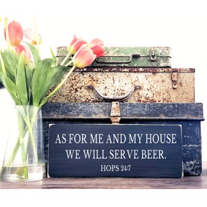 Puede incluir: Un letrero de madera negro con texto blanco que dice "As for me and my house we will serve beer. Hops 24:7". El letrero est&aacute; colocado sobre tres maletas de metal vintage.