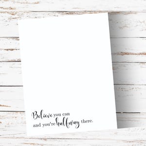 May include: A white paper print with the text "Believe you can and you're halfway there" in black script. The print is angled against a white wooden background.