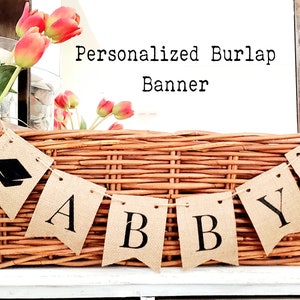 May include: A burlap banner with the name "Abby" spelled out in black letters. Each letter is on a separate flag with a graduation cap on the end flags. The banner is hanging over a wicker basket.