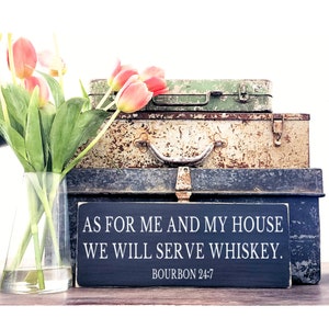 以下が含まれることがあります： 「As for me and my house we will serve whiskey. Bourbon 24:7」と白文字で書かれた黒い木製の看板。看板は3つのビンテージメタルボックスの上に置かれています。