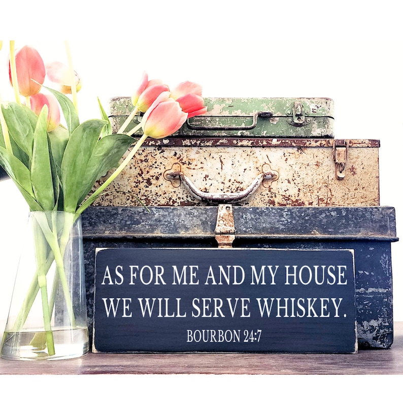 以下が含まれることがあります： 「As for me and my house we will serve whiskey. Bourbon 24:7」と白文字で書かれた黒い木製の看板。看板は3つのビンテージメタルボックスの上に置かれています。