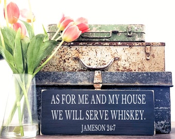 Op de afbeelding: Een zwart houten bord met witte tekst die luidt "As for me and my house we will serve whiskey. Jameson 24:7". Het bord ligt op drie vintage metalen koffers.