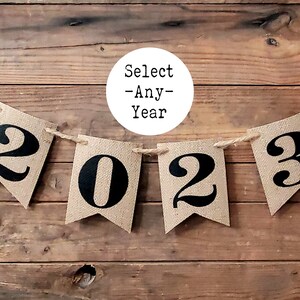 May include: A burlap banner with black graduation caps and the year 2023 printed on it. The banner is strung with twine and hangs on a wooden background. A white circle with the text "Select -Any- Year" is in the background.