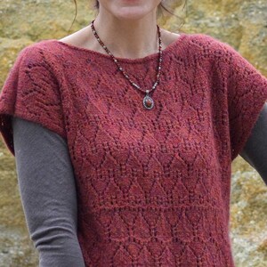 Puede incluir: Una mujer lleva una blusa de ganchillo de color granate con un collar de cuentas y un colgante. La blusa tiene un dobladillo festoneado y mangas cortas.