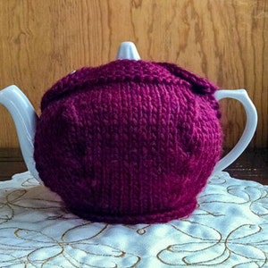 Puede incluir: Una tetera blanca con un calienta-tetera de punto burdeos. El calienta-tetera es un punto sencillo con un patrón texturizado.