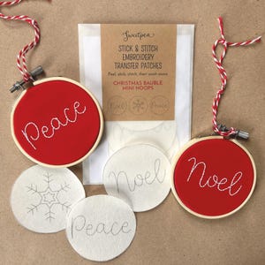May include: Two round wooden embroidery hoops containing red fabric with white stitched text reading Peace and Noel, each with red and white twine. Includes a package of Sweetpea Stick & Stitch embroidery transfer patches with snowflake and text designs.