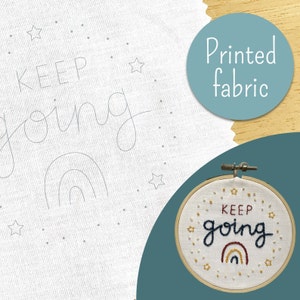 May include: A white fabric with a printed design of a rainbow and the text "KEEP going" surrounded by stars. The fabric is in a hoop and the text is embroidered in colourful thread.