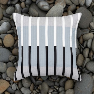 May include: A gray and black striped pillow with a geometric pattern. The pillow is made of a soft, textured fabric and is sitting on a bed of gray rocks.