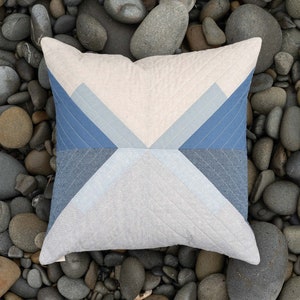 May include: A square pillow with a geometric pattern of blue and white triangles on a beige background. The pillow is lying on a bed of smooth, gray river rocks.