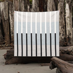 May include: A black, white, and gray striped quilt hanging in front of a weathered wooden background. The quilt is made of fabric with a subtle texture.