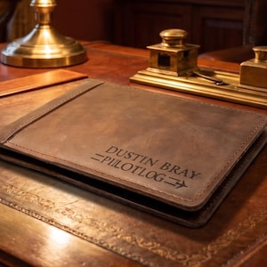 May include: A dark brown leather pilot logbook cover with visible stitching along the edges. The front cover is embossed with the text DUSTIN BRAY and PILOTLOG, accompanied by a small aeroplane icon flanked by two horizontal lines.
