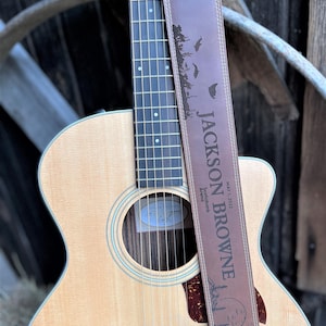 Peut inclure: Guitare acoustique avec une sangle en cuir marron. La sangle porte en relief le nom "JACKSON BROWNE", une date et une scène de nature. La guitare a une finition en bois clair et une touche sombre.